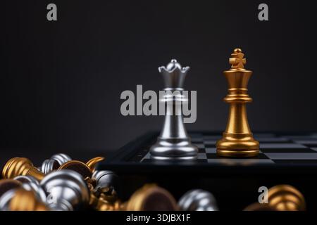 goldener König und silberne Königin umgeben von silbernen und goldenen Schachstücken, die hinter dem Schachbrett liegen. Stockfoto
