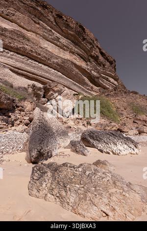 Ein beeindruckendes Bild einer massiven, mehrschichtigen Klippe, die einen abgelegenen Strand auf der Arabischen Halbinsel in Oman dominiert. Große, strukturierte Felsbrocken sind verstreut Stockfoto