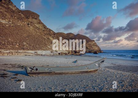 Ein traditionelles, altes omanisches Fischerboot (Dhow), bemalt mit lokalen Motiven, ruht an einem Sandstrand mit kleinen Muscheln. Im Hintergrund, robust, A Stockfoto
