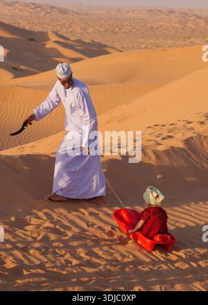 Ein omanischer Mann in traditioneller Kleidung zieht ein Kind auf einem roten Schlitten durch weite, sonnendurchflutete Wüstendünen. Diese herzerwärmende Szene vermittelt Familienspaß und trave Stockfoto