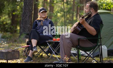 Ein paar Camper sitzen in der Nähe eines Zeltes, während sie im Wald campen und Gitarre spielen. Familiencamping. Stockfoto