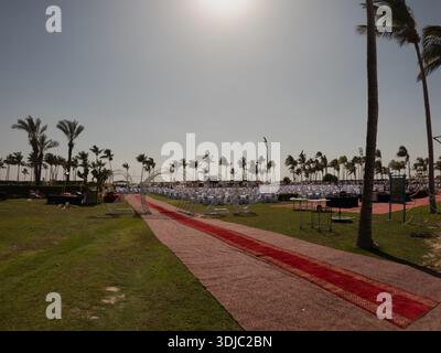 Ein Veranstaltungsraum im Freien in Salalah, Oman, mit einem roten Teppich, der zu einer Reihe von weißen Tischen und Stühlen unter Palmen führt. Ideal für den Transport Stockfoto