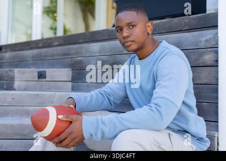 Afroamerikaner sitzt auf einer Holzbank am Eingang des modernen Hauses und hält rot-weißen Fußball Stockfoto