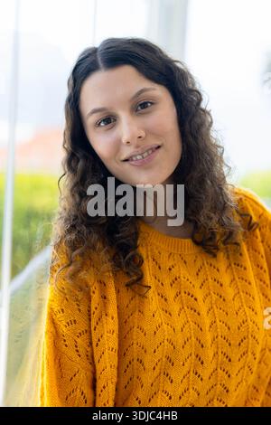 Frau in ihren 20ern, die sich nahe eines großen Glasfensters lehnte und einen senfgelben Strickpullover trug Stockfoto