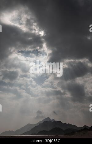 Eine vertikale Aufnahme, die Sonnenstrahlen einfängt, die durch dramatische, dunkle Wolken über einer Linie von Silhouettenbergen in Sadah, Oman, brechen. Die Stimmung ist mächtig und Stockfoto