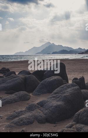 Eine ruhige, stimmungsvolle Landschaftsaufnahme eines felsigen Sandstrandes in Sadah, Oman. Große, dunkle Felsbrocken sind im Vordergrund verstreut und führen zu sanften Formen Stockfoto