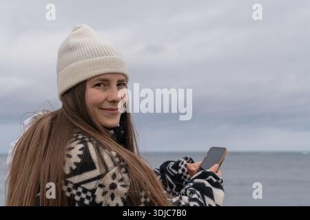 Frau lächelt und hält Smartphone am bewölkten Tag am Meer Stockfoto