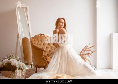 Frau in einem fließenden weißen Kleid sitzt auf einem Vintage-Sessel mit Spiegel Stockfoto