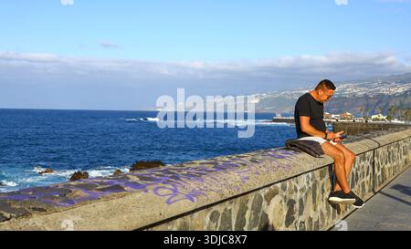 Mann, der auf einer Steinmauer sitzt und den Ozean überblickt, mit Mobilgerät und Küstenstadt im Hintergrund Stockfoto