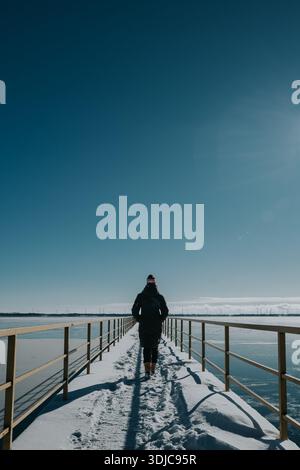 Frau in Winterkleidung, die am Wintertag auf einem verschneiten Pier weggeht. Stockfoto
