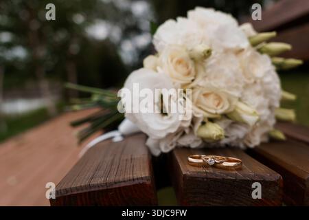 Eine Nahaufnahme eines romantischen weißen Hochzeitsstraußes mit Rosen und Lisianthus, der neben goldenen Eheringen auf einer rustikalen Holzbank liegt und eine ruhige Atmosphäre einnimmt Stockfoto