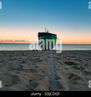 Ein verlassenes, verwittertes grünes Fischerboot sitzt am Strand, während die Sonne untergeht. Der Himmel wechselt von tiefem Blau zu sanftem Orange und rosa h Stockfoto
