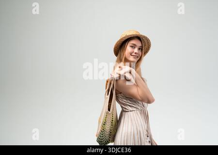 Die lächelnde junge Frau trägt einen Strohhut und ein leichtes Sommerkleid und hält eine wiederverwendbare Einkaufstasche auf neutralem Hintergrund. Image steht für nachhaltige Geschäfte Stockfoto