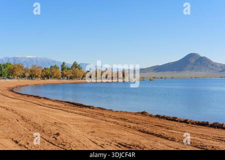 Das ruhige Wasser des Perris-Sees reflektiert einen klaren blauen Himmel, der von einem geschwungenen Sandstrand, farbenfrohen Herbstbäumen und majestätischen, fernen Bergen umgeben ist. Stockfoto