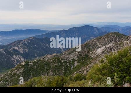 Weitläufiger Panoramablick auf mehrschichtige blaue Bergrücken unter einem bewölkten Himmel in den San Gabriel Mountains, mit einer trüben Stadt in der Ferne Stockfoto