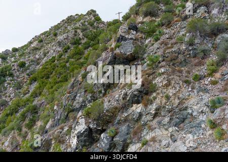Detaillierte Aufnahme einer steilen, felsigen Klippe in den San Gabriel Mountains, die abwechslungsreiche Geologie, karges Kaparral und einen einsamen hölzernen Strommast auf der Spitze zeigt Stockfoto