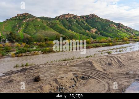 Der trübe Fluss fließt vorbei an lebhaften grünen, von Felsen übersäten Hängen des Mt. Rubidoux unter einem bewölkten Himmel, Reifenspuren am Sandufer darunter. Stockfoto