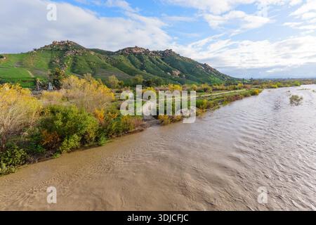 Ein kräftiger, schlammiger Fluss fließt vorbei an lebendiger Ufervegetation und einem unbefestigten Pfad mit grünen, felsigen Hügeln unter einem dynamischen, teils bewölkten Himmel. Stockfoto