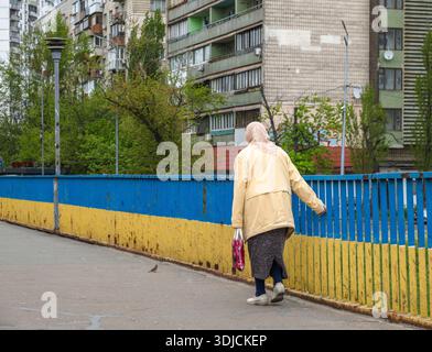 Kiew, Ukraine - 7. Mai 2022: Ältere Frau im Kopftuch geht entlang der Kiewer Brücke mit blauem und gelbem Geländer, symbolisiert ukrainische Nationalfarben, Widerstandsfähigkeit und Hoffnung in der Ukraine Stockfoto