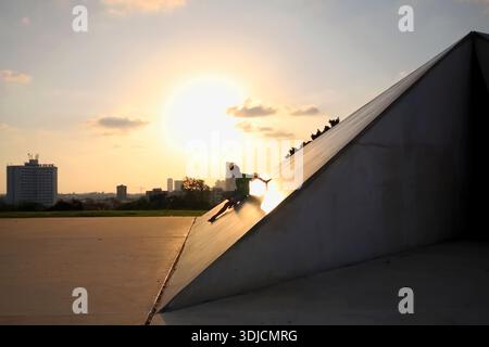 Die Silhouette eines Kindes auf einer Betonpyramidenskulptur bei Sonnenuntergang, eingerahmt von einer massiven glühenden Sonne und der trüben Skyline der Stadt Tel Aviv in der Ferne. Stockfoto