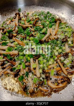 Nahaufnahme von Shimeji-Pilzen, die mit gehackten grünen Zwiebeln in einer Edelstahlpfanne anbraten. Pilze, die in heißem Öl mit sichtbaren Blasen braten, aufgefangen Stockfoto