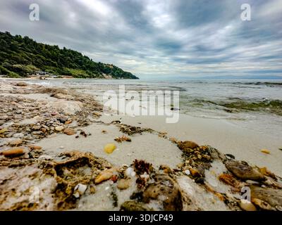 Weite Aussicht auf eine felsige Mittelmeerküste in der Nähe von Afytos Dorf in Chalkidiki, Griechenland. Kieselsteine, flaches Wasser, sanfte Wellen und ferne Hügel unter A Stockfoto