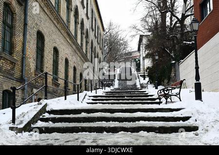 Außentreppe im Winter. Schneebedeckte Steintreppen. Schwindende perspektivische Ansicht. Graues Metallrohrgeländer. Braune Holz- und Eisenbank. Backsteinbau Stockfoto
