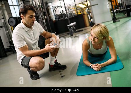 Kaukasischer junger erwachsener Mann kniend neben kaukasischer junger erwachsener Frau, die Plank-Übungen auf Matte im Fitnessstudio durchführt, Mann-Timing-Sitzung mit Stoppuhr, Physiotherapie-Rehabilitation Stockfoto