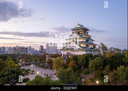 Osaka, Japan - 7. November 2023: Der Hauptturm der Burg Osaka wird in der Abenddämmerung beleuchtet, mit einem umliegenden platz, Bäumen und modernen Stadtgebäuden, die im Hintergrund unter einem teilweise bewölkten Himmel zu sehen sind. Stockfoto