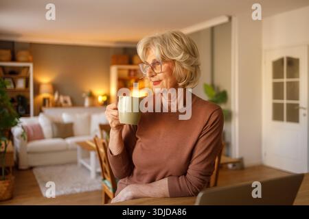 Eine aufmerksame Seniorin mit Brille blickt weg und trinkt Kaffee, während sie sich morgens zu Hause entspannt Stockfoto