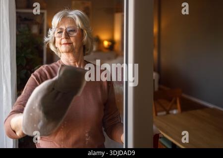 Porträt einer selbstbewussten älteren Frau mit kurzen Haaren und Brille, die zu Hause nasse Glasfenster mit Serviette abwischt Stockfoto