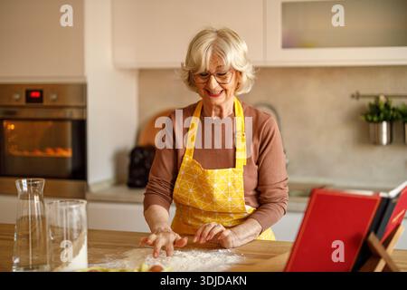 Lächelnde Seniorin in Schürze, die Ei in Mehl auf der Kücheninsel mischt, während sie Teig für hausgemachte Kekse zubereitete Stockfoto