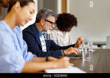 Konzentrierter Geschäftsmann, der über einem Laptop arbeitet, während weibliche Kollegen im Besprechungsraum Dokumente gemeinsam analysieren Stockfoto