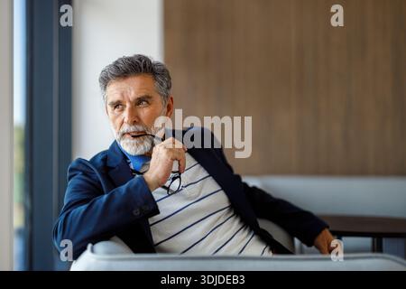 Porträt eines gutaussehenden, bärtigen Geschäftsmannes, der eine Brille hält und nachdenklich wegschaut, während er im Büro auf dem Sofa sitzt Stockfoto