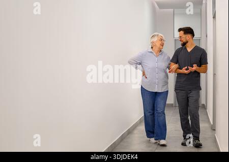 Volle Länge der älteren Frau, die mit dem männlichen Arzt Arm in Arm geht, während sie Symptome und Behandlung in der Krankenhaus-Lobby bespricht Stockfoto