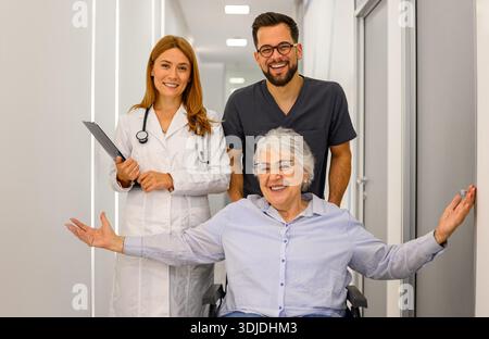 Porträt selbstbewusster Ärzte, die fröhliche Senioren im Rollstuhl entlang des Krankenhauskorridors ziehen Stockfoto