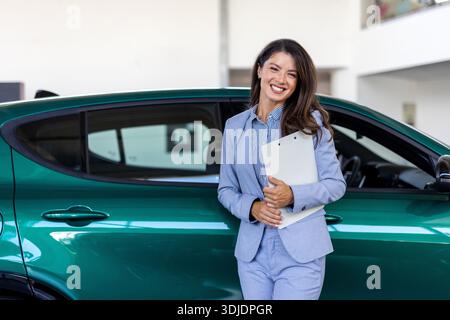 Lächelnde Verkäuferin, die einen Tablet-PC hält, während sie die Kamera im Ausstellungsraum eines neuen Autos betrachtet. Professioneller Autohändler posiert stolz im Autohaus und lächelt zu Stockfoto
