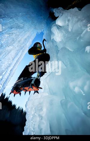 Ein Mann in gelber Jacke klettert mit Eisaxt und Steigeisen an einer gefrorenen Eiswand am großen Wasserfall gegen blaue Ski in den Winterbergen. Sport Bergsteiger Stockfoto