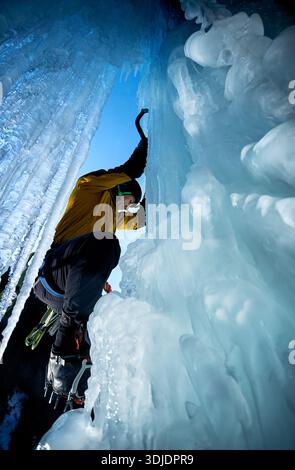 Ein Mann in gelber Jacke klettert mit Eisaxt und Steigeisen an einer gefrorenen Eiswand am großen Wasserfall gegen blaue Ski in den Winterbergen. Sport Bergsteiger Stockfoto
