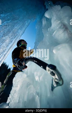 Ein Mann in gelber Jacke klettert mit Eisaxt und Steigeisen an einer gefrorenen Eiswand am großen Wasserfall gegen blaue Ski in den Winterbergen. Sport Bergsteiger Stockfoto