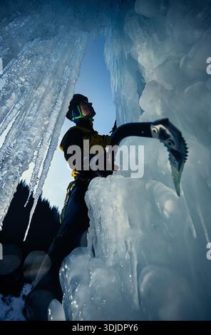 Ein Mann in gelber Jacke klettert mit Eisaxt und Steigeisen an einer gefrorenen Eiswand am großen Wasserfall gegen blaue Ski in den Winterbergen. Sport Bergsteiger Stockfoto