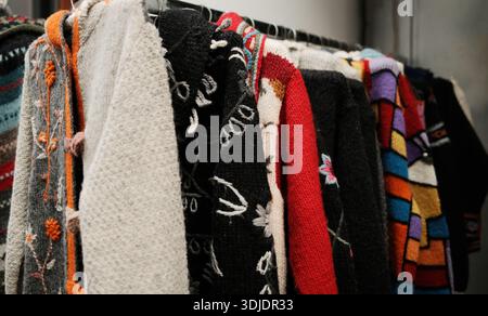 Nahaufnahme handgefertigter Strickpullover und Tücher, die an einem Gestell hängen und kräftige Texturen, buntes Garn und traditionelle Muster zeigen Stockfoto
