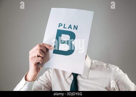 Geschäftsmann hält ein weißes Papierschild mit PLAN B-Text vor seinem Gesicht für Backup-Strategie, alternative Lösung und Verfügbarkeitsplan. Selektiv Stockfoto