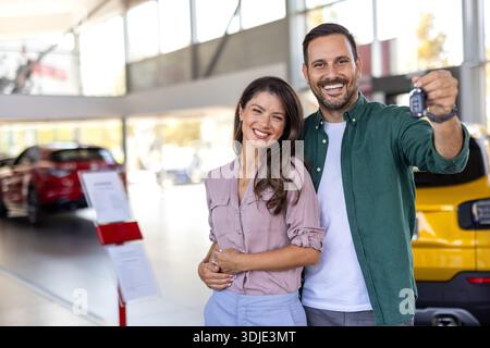 Glückliches, schönes, liebevolles Paar, das im Salon ein neues Auto kauft, über Luxusauto steht, sich umarmt und den Schlüssel aus ihrem neuen Auto zeigt, Kopierraum Stockfoto