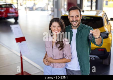 Glückliches, schönes, liebevolles Paar, das im Salon ein neues Auto kauft, über Luxusauto steht, sich umarmt und den Schlüssel aus ihrem neuen Auto zeigt, Kopierraum Stockfoto