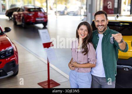 Glückliches, schönes, liebevolles Paar, das im Salon ein neues Auto kauft, über Luxusauto steht, sich umarmt und den Schlüssel aus ihrem neuen Auto zeigt, Kopierraum Stockfoto