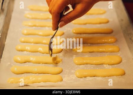 Roher Teig für Savoyardi-Kekse zur Herstellung von Tiramisu-Kuchen liegt auf Pergamentpapier. Die Hand einer Frau verteilt den Teig mit einem Löffel Stockfoto