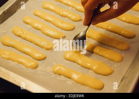 Roher Teig für Savoyardi-Kekse zur Herstellung von Tiramisu-Kuchen liegt auf Pergamentpapier. Die Hand einer Frau verteilt den Teig mit einem Löffel Stockfoto