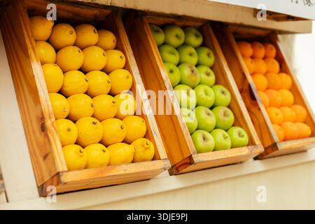 Helle Zitronen, grüne Äpfel und Orangen sind ordentlich auf Holztabletts angeordnet. Die Marktkulisse ist voller Besucher und die Sonne scheint auf Stockfoto