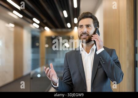 Bärtiger männlicher Profi im Anzug, der sich auf seinem Mobiltelefon unterhalten und mit einem Kunden oder Kollegen kommunizieren kann, während er durch ein modernes Büro geht Stockfoto